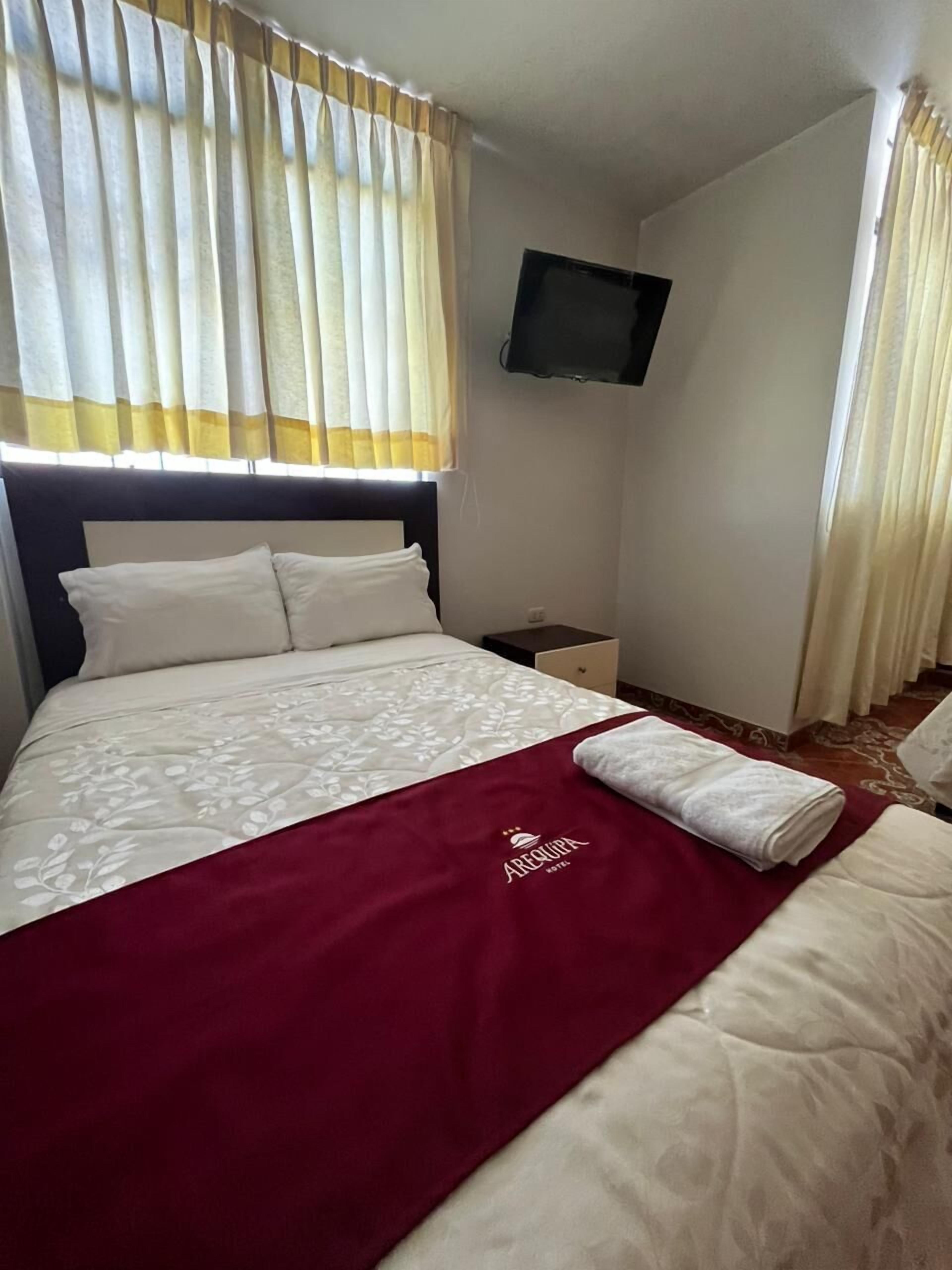 A neatly made bed with a red blanket and a white pillow, a nightstand with a lamp, and a television mounted on the wall.