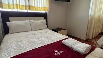 A neatly made bed with a red blanket and a white pillow, a nightstand with a lamp, and a television mounted on the wall.