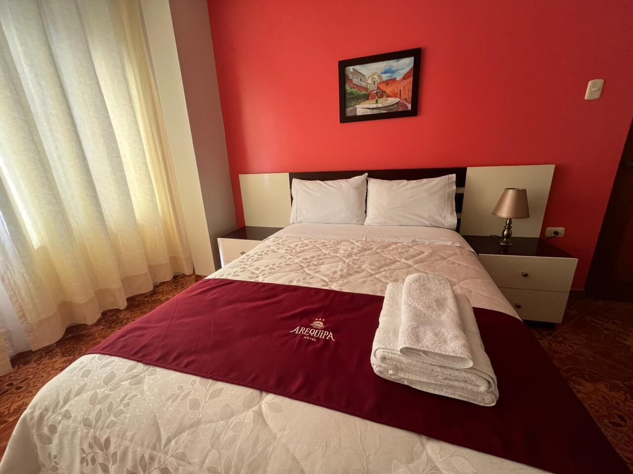 Una habitación de hotel con cama, mesita de noche, lámpara, un cuadro en la pared y una ventana con cortinas.