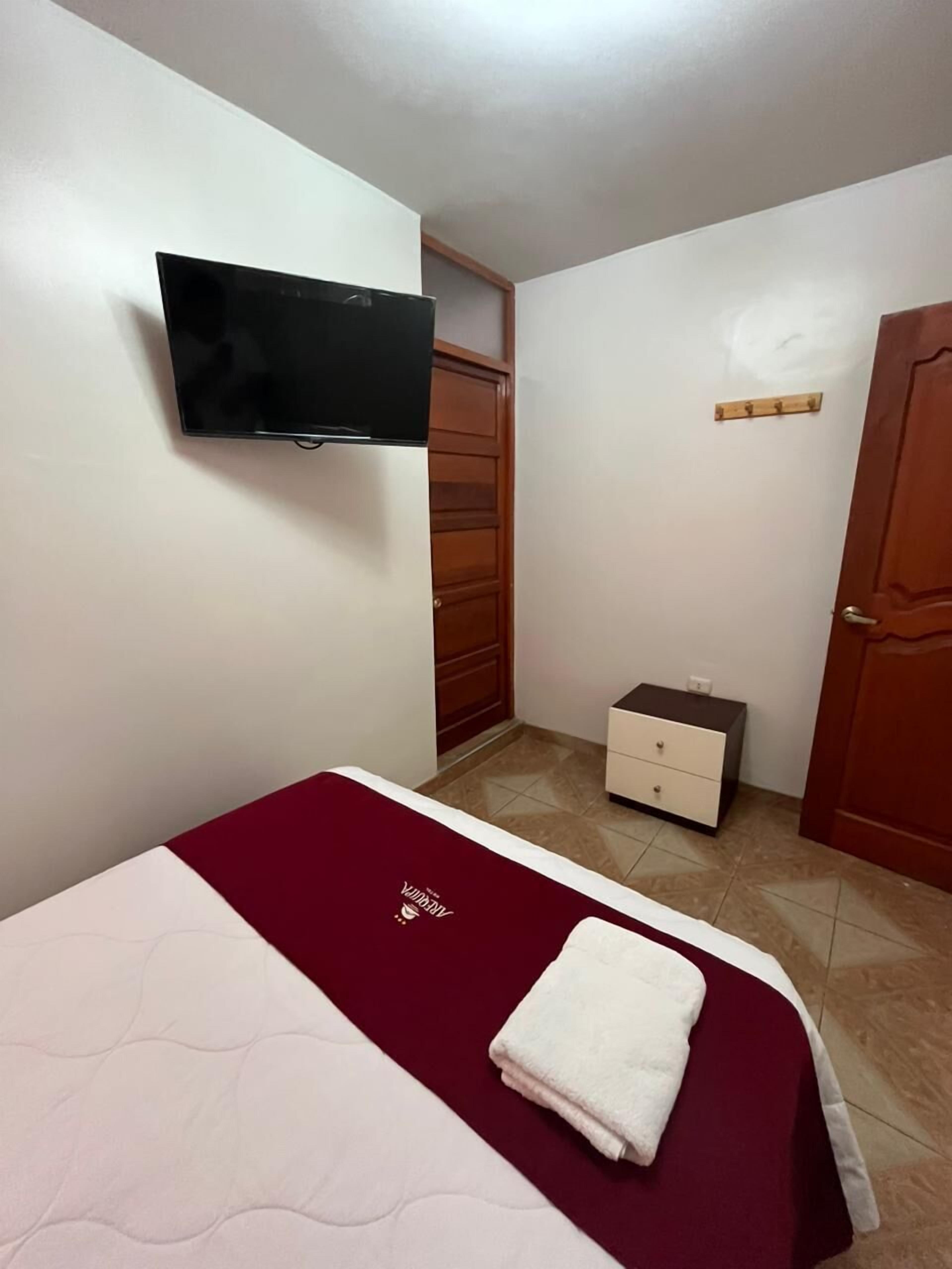 Un dormitorio pequeño con una cama, un televisor montado en la pared, un armario de madera, una mesita de noche y una puerta.