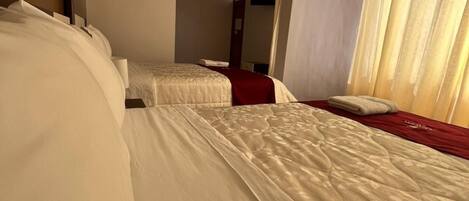 Habitación de hotel con dos camas, cada una con ropa blanca y un detalle rojo en la cabecera, y una ventana con cortinas translúcidas.