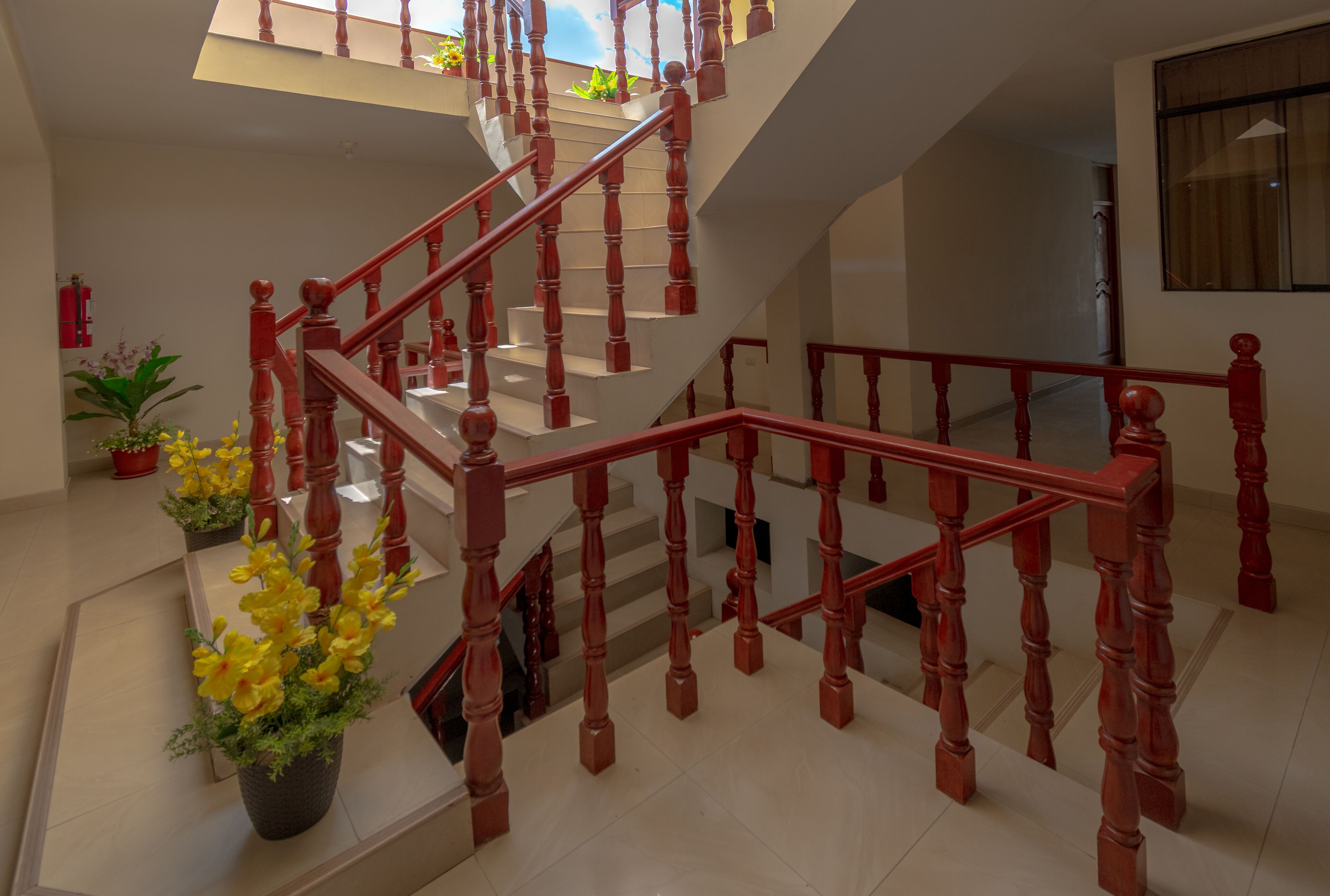 Una escalera de madera de varios niveles con barandilla roja, rodeada de plantas en macetas y un suelo de baldosas.