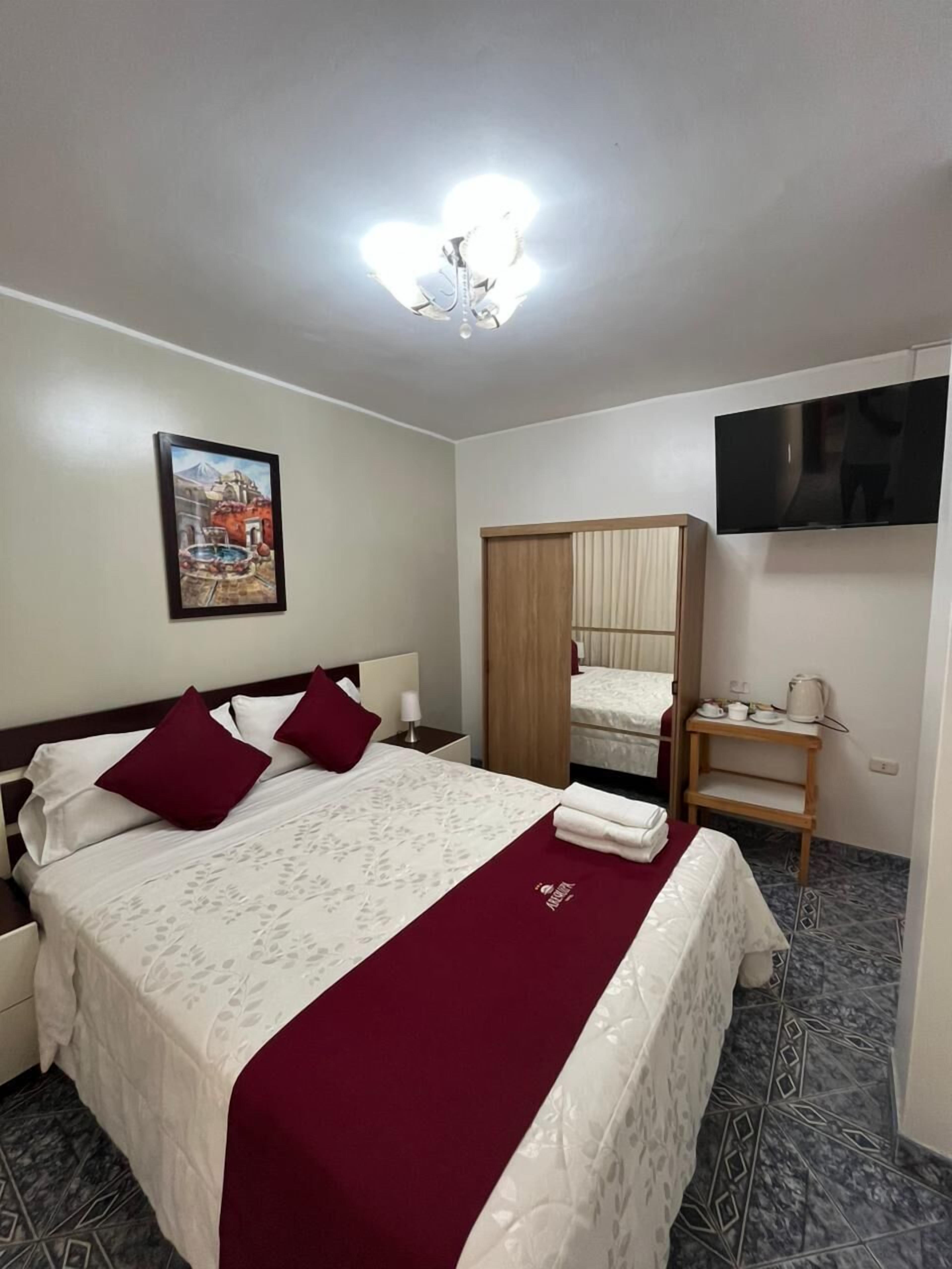 Habitación de hotel con cama, televisor, armario de madera, una mesita con tazas y un hervidor, y un cuadro en la pared.