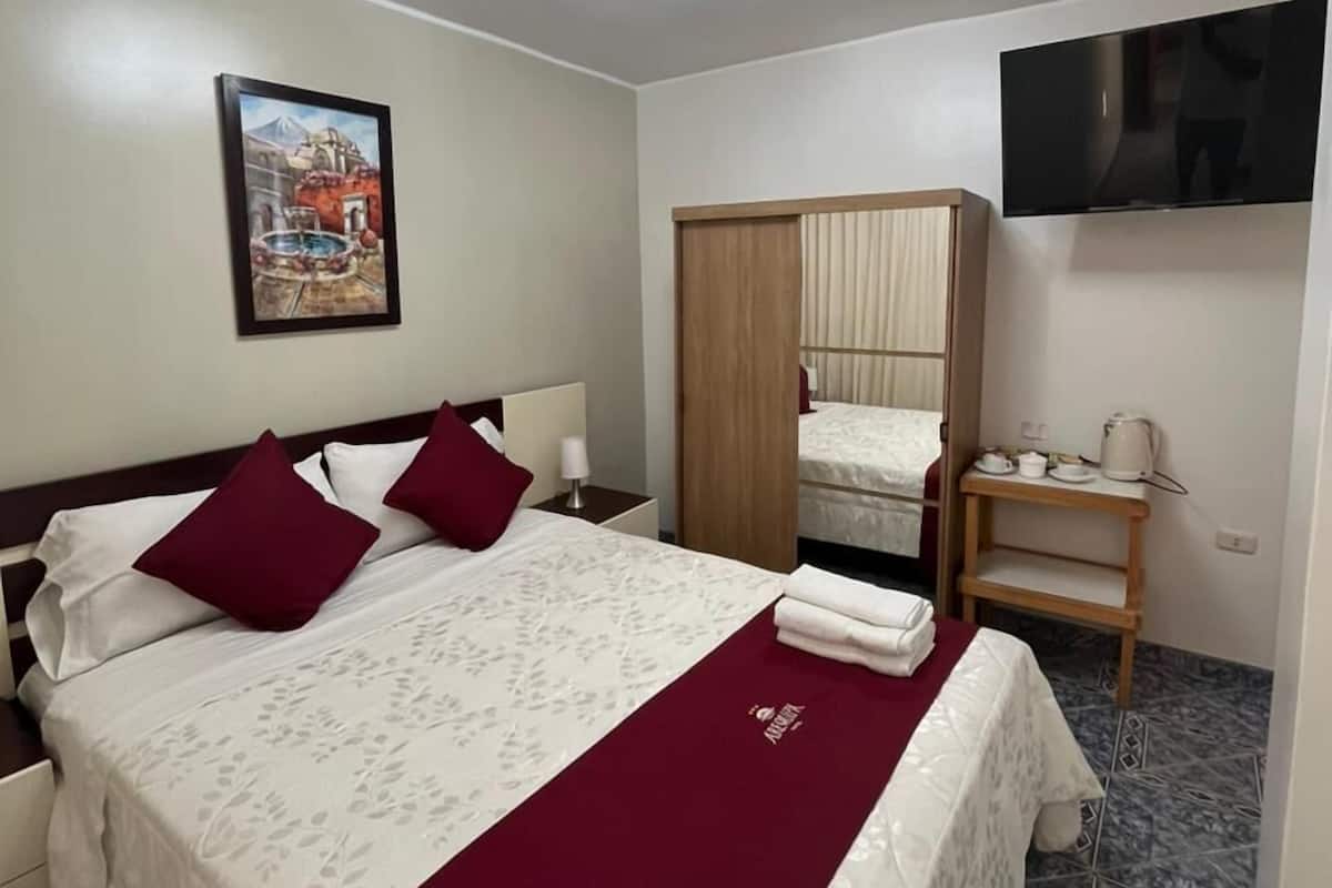 A hotel room with a bed, a TV, a wooden wardrobe, a small table with cups and a kettle, and a painting on the wall.