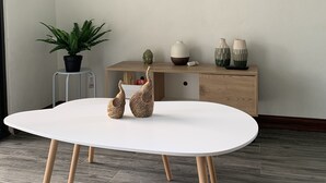 Een moderne woonkamer met een witte tafel, twee decoratieve keramieken beeldjes, een wandgemonteerde flatscreen-tv, een houten kast met decoratieve objecten en een potplant.