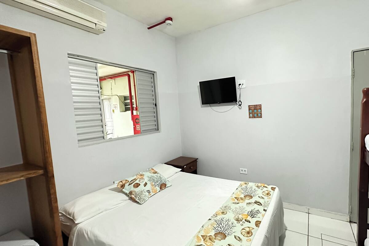 A bedroom with a bunk bed, a single bed with a patterned bedspread, a television on the wall, and a window with blinds.