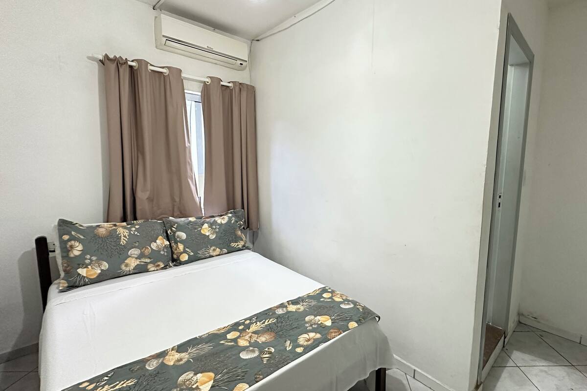 A small, neatly arranged bedroom with a bed, curtains, an air conditioning unit, and a door.
