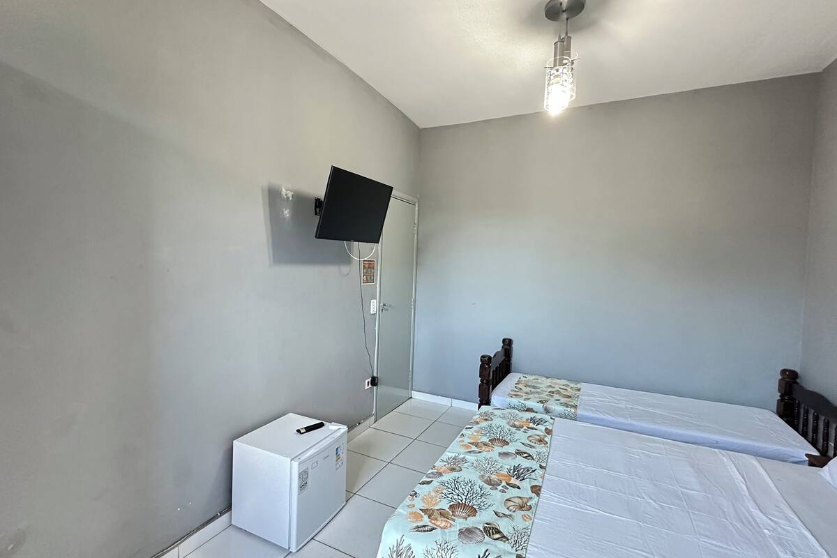 A bedroom with a bed, a TV mounted on the wall, a small white cabinet, and a ceiling light fixture.