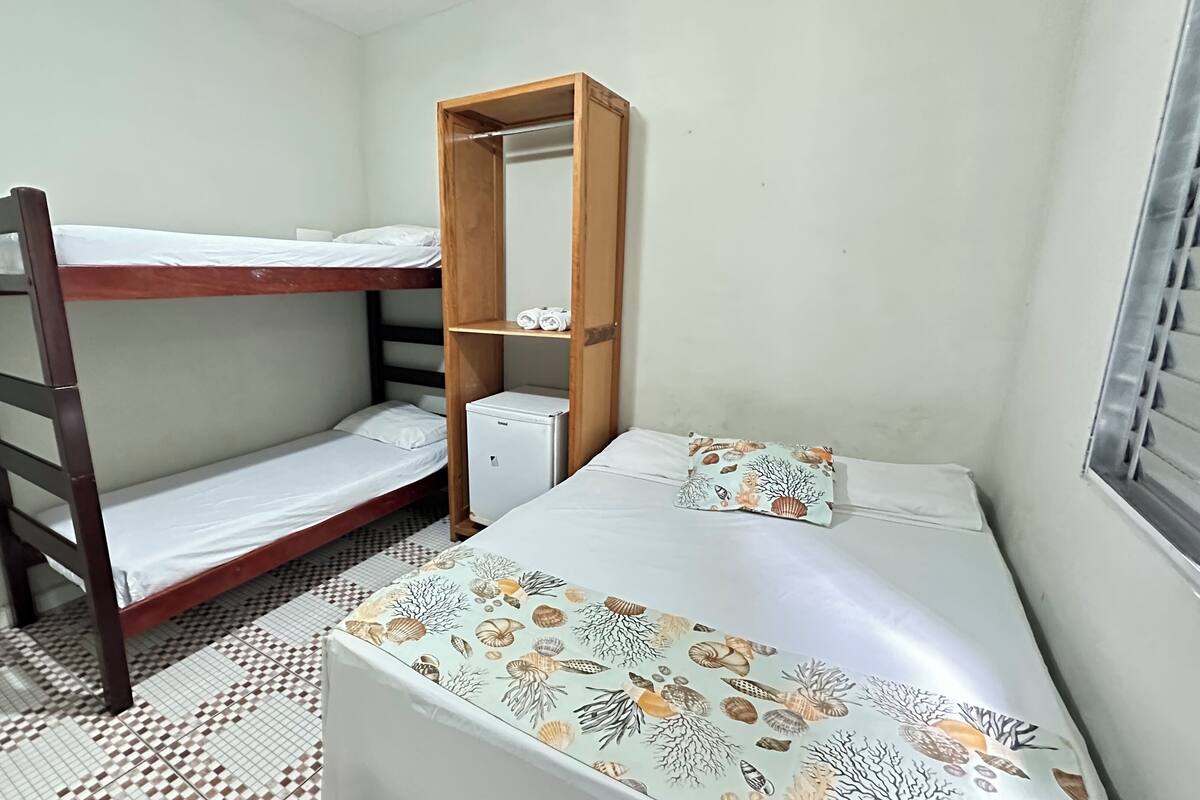A small, compact room with a bunk bed, a single bed with a patterned bedspread, a small refrigerator, and a window with blinds.