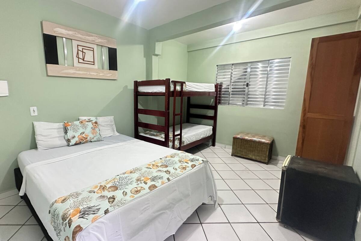 A room with a bunk bed, a single bed, a small table, and a window with blinds.