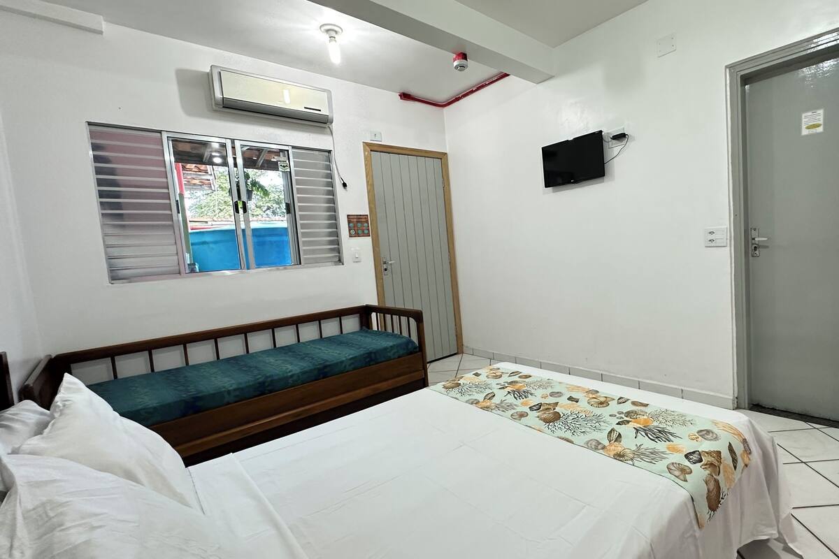 A bedroom with a bed, a bench, a window with blinds, an air conditioner, a television, and a door.
