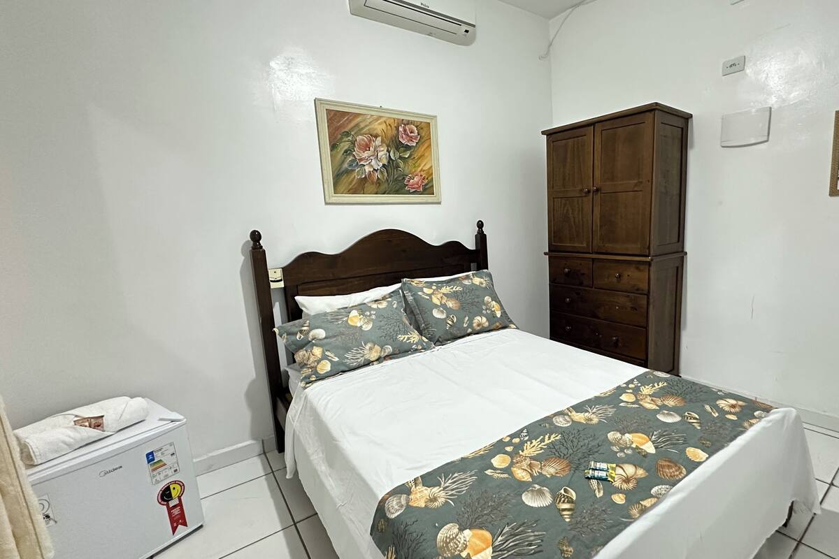 A bedroom with a bed, a wardrobe, a painting on the wall, and a small air conditioning unit.