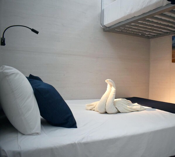 Una cama individual con un colchón blanco, una almohada azul oscuro y una toalla blanca doblada encima. Una lámpara negra está montada en la pared a la izquierda.