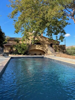 Un edificio de piedra con piscina, un árbol y un cielo azul despejado.