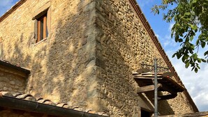 Un bâtiment en pierre doté d’une terrasse, de deux chaises en métal, d’une table et d’une porte en bois.