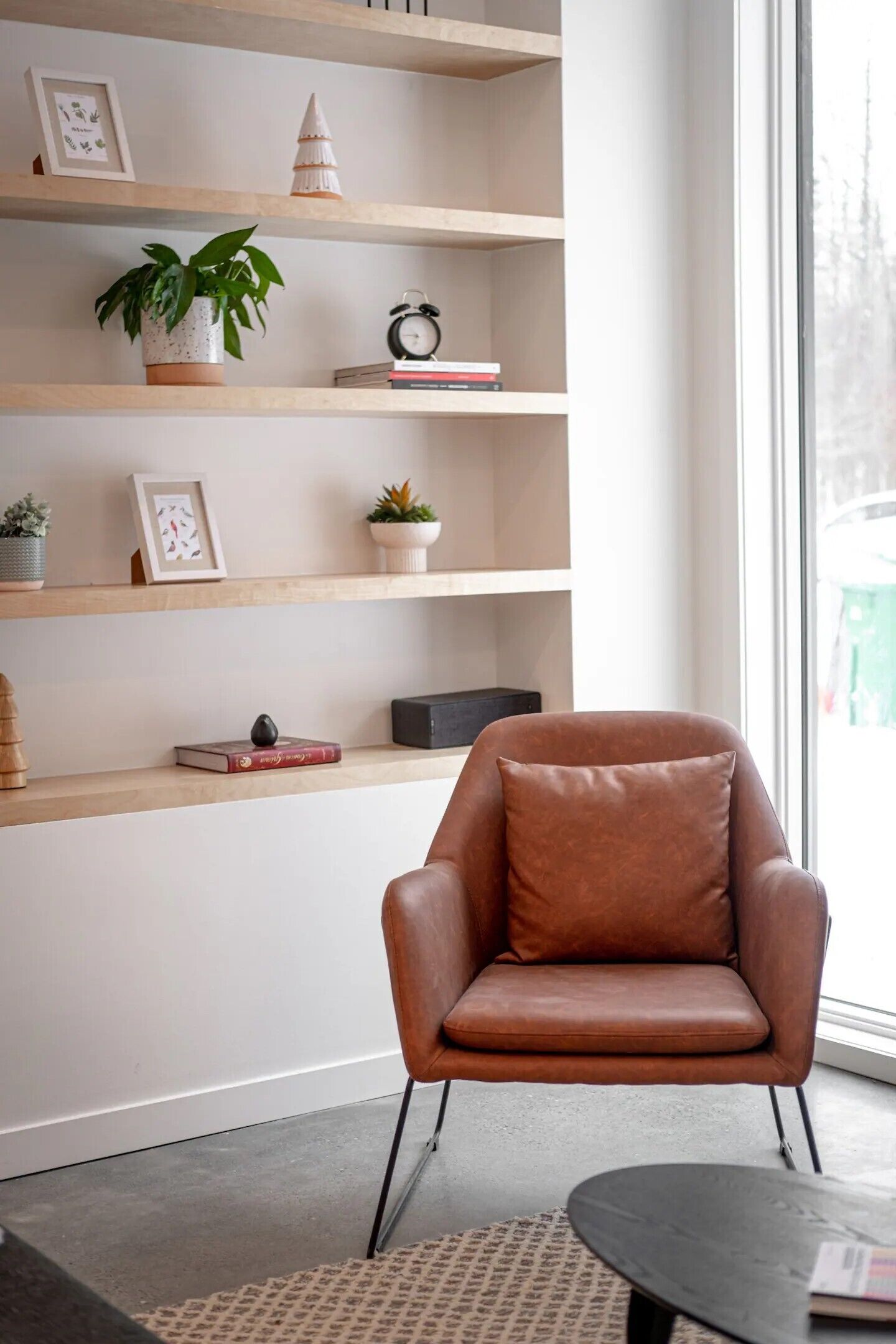 A modern living room with a brown leather armchair, wooden shelves with decorative items, and a large window.