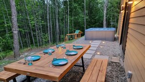 Un'area pranzo all'aperto con tavolo in legno apparecchiato per quattro persone, circondata da ghiaia, con una vasca idromassaggio e un molo in legno sullo sfondo.