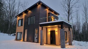 Une maison moderne à deux étages, dotée d’un porche abrité, entourée de neige et d’arbres.