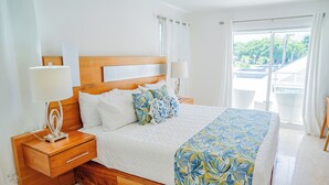 Een slaapkamer met een houten hoofdbord, een bed met wit beddengoed en een bloemenbedekking in blauw en geel, twee bedlampjes en uitzicht op een balkon met witte rieten meubels.
