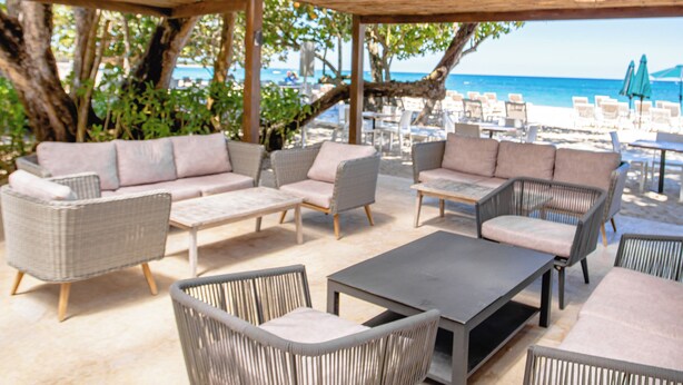 Een terras buiten met rieten meubilair en een houten pergola, met uitzicht op een strand.