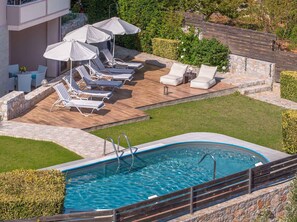 A swimming pool with lounge chairs and umbrellas, surrounded by a stone deck and a grassy area. - Villa Stefania (Chania)