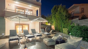 A well-lit outdoor seating area with cushioned chairs and a table, surrounded by a stone wall and greenery. - Villa Stefania (Chania)