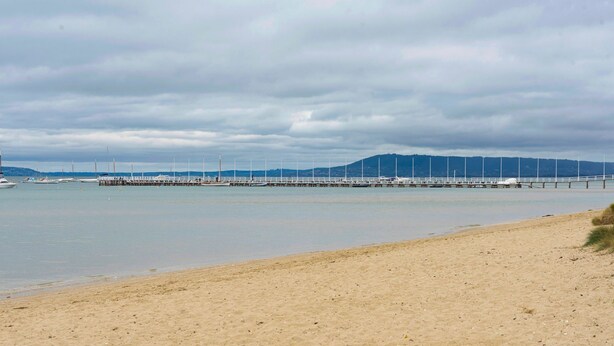 Una playa con un muelle que se adentra en aguas tranquilas, una orilla arenosa y colinas al fondo.