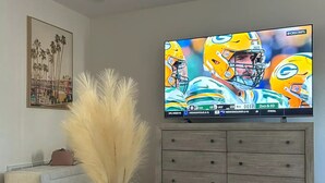 Una sala de estar con televisión de pantalla plana, una mesa de centro de vidrio, un sofá, un jarrón decorativo y un mueble de madera.