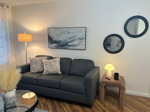 A living room with a grey sofa, two round mirrors, a framed abstract painting, a floor lamp, and a small side table with a lamp.