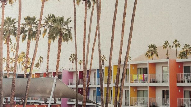 Un hotel frente a la playa con edificios de varios pisos, palmeras y un área de piscina con tumbonas y sombrillas.