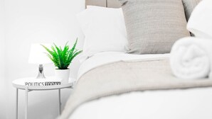 Una cama bien hecha con cabecera gris, almohada blanca y almohada gris a rayas. Hay una mesita de noche pequeña de color blanco con una lámpara, una planta en maceta y un libro titulado 'POLITICS TODAY'.