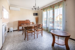A dining area with a wooden table and chairs, a small round table, a chandelier, a television mounted on the wall, and a window with floral curtains.