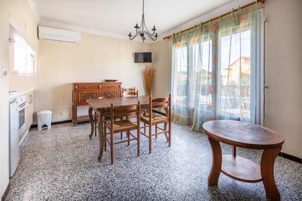 A dining area with a wooden table and chairs, a small round table, a chandelier, a television mounted on the wall, and a window with floral curtains.