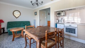 Un espace de vie compact comprenant une table à manger, des chaises, un canapé vert, un petit coin cuisine avec un micro-ondes et un four, et un lustre.