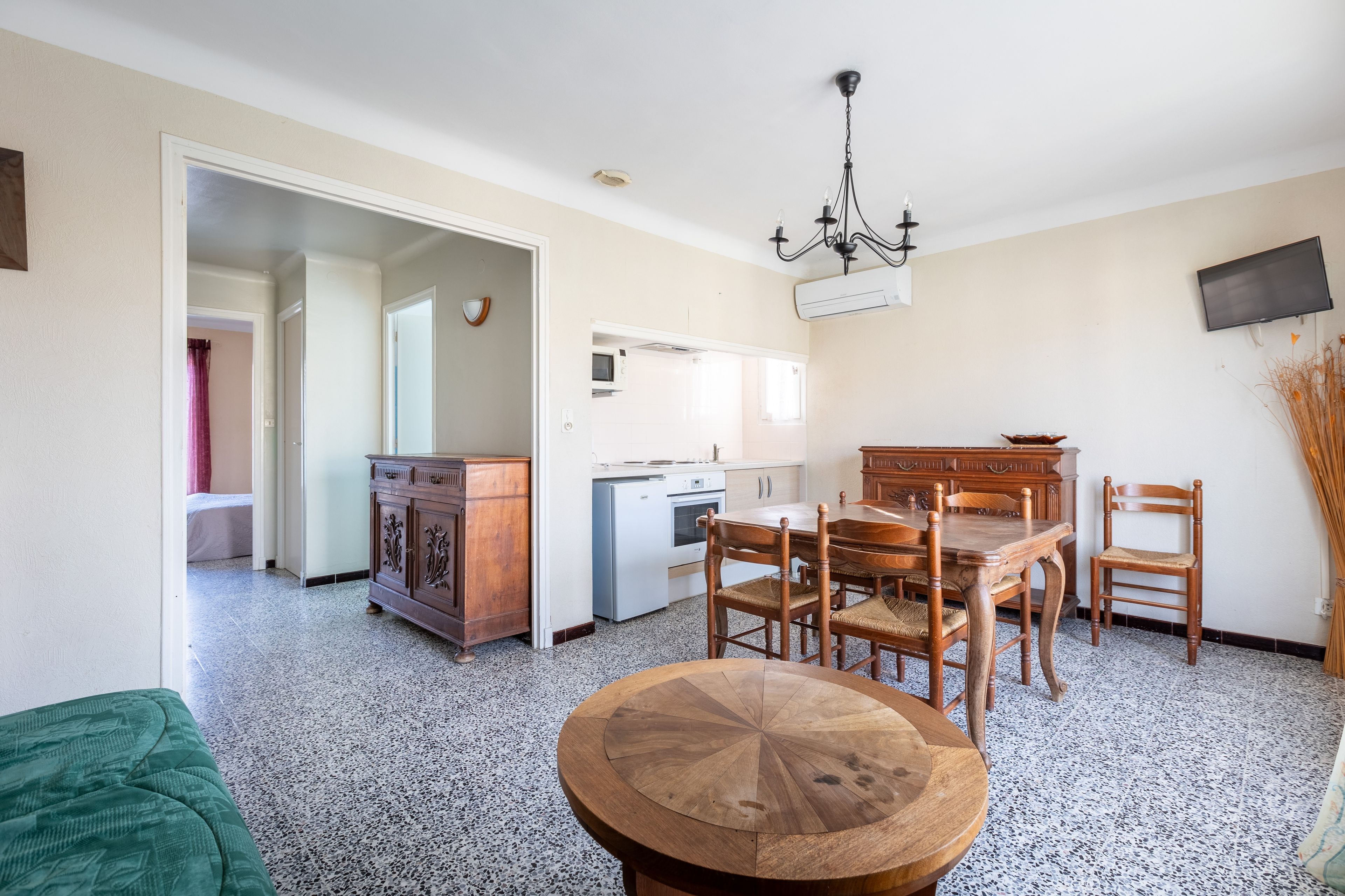 Uma área de refeições com uma mesa de madeira redonda, cadeiras, cozinha com micro-ondas e televisão fixada na parede.