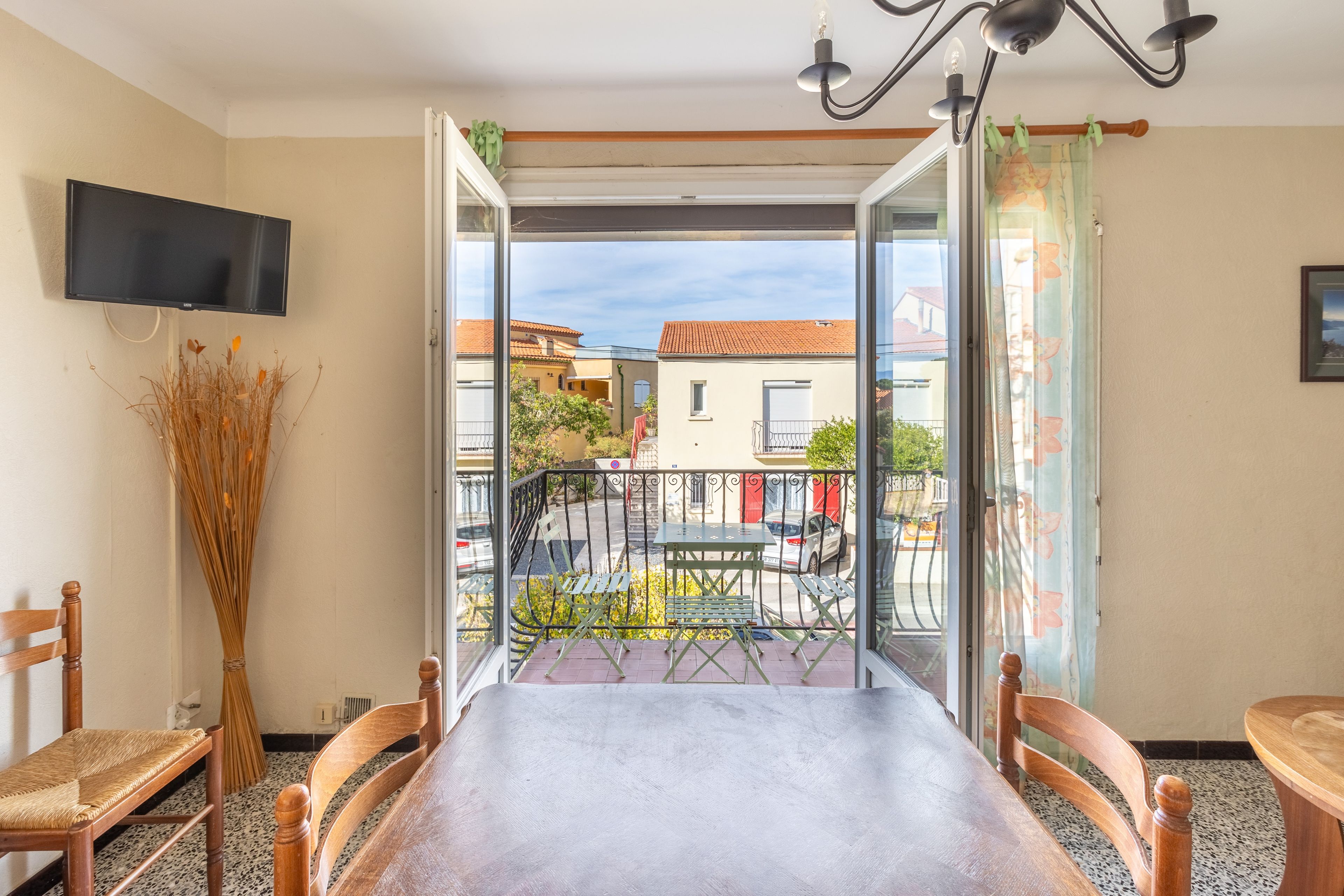 A room with a balcony, a dining table, chairs, a TV, and a vase with dried plants.
