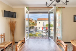 A room with a balcony, a dining table, chairs, a TV, and a vase with dried plants.