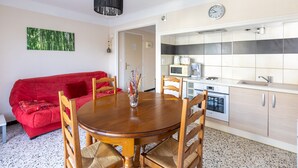 A compact living space with a dining area, a red sofa, wooden chairs, a kitchen with a microwave, and a clock on the wall.