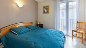 Ein Schlafzimmer mit Bett, Nachttisch, Stuhl, einem Gemälde und einem Balkon mit Vorhängen.