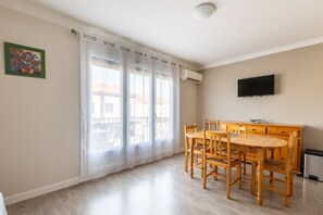 A room with a dining table, chairs, a TV mounted on the wall, and an air conditioning unit. - Mandarine - Appt à 10min d'Argelès-Sur-Mer (Saint-André)