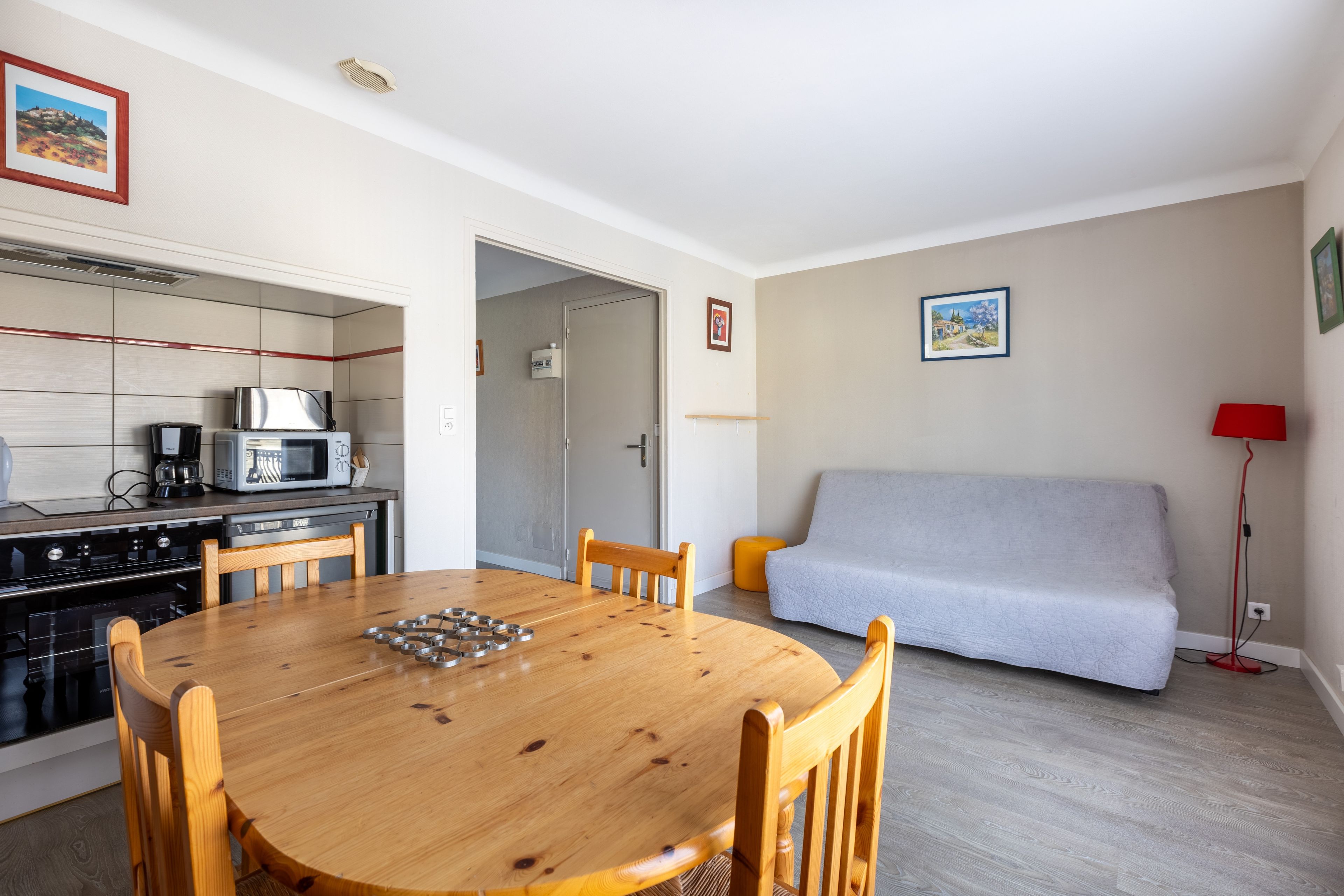 Een ruimtelijk ingerichte leefruimte met een eettafel, stoelen, een kitchenette, een bank en een rode vloerlamp.