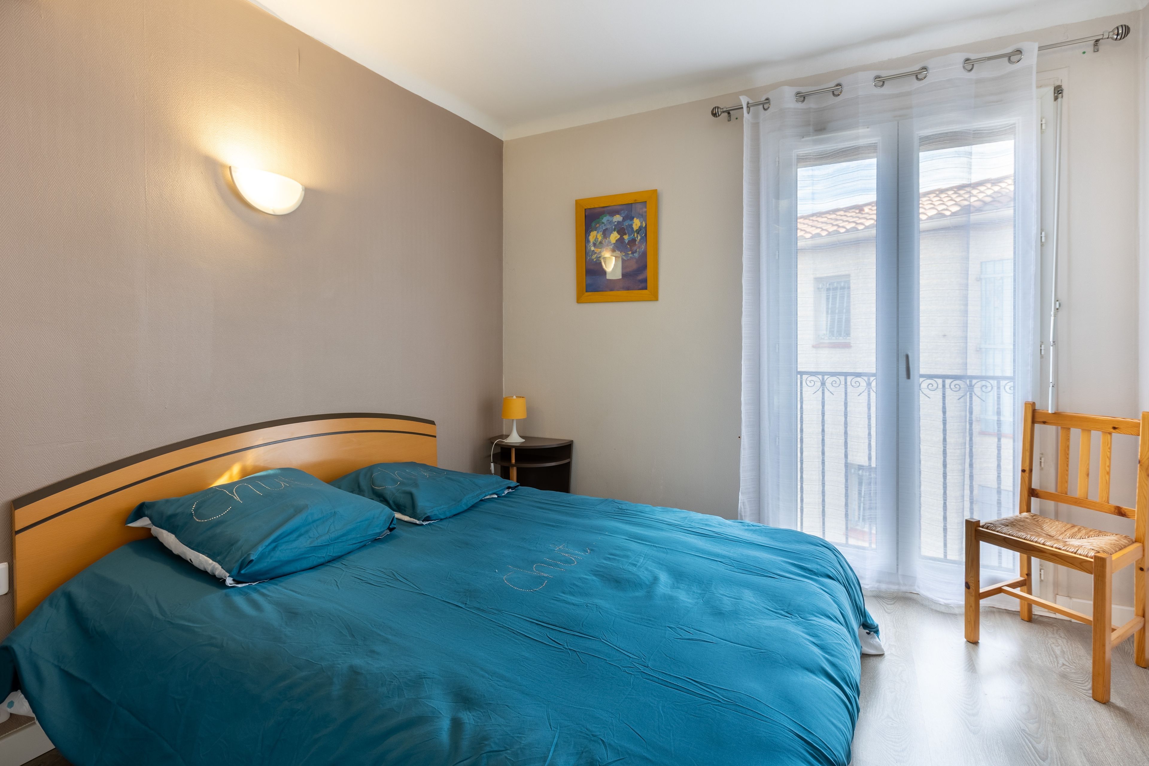 Une chambre à coucher avec un lit, une table de chevet, une chaise, une fenêtre avec des rideaux et un tableau au mur.