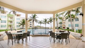 Un espacio al aire libre techado con piscina, mesas y sillas para comer, rodeado de palmeras y vistas a un edificio con balcones.
