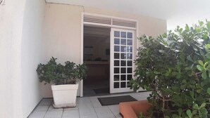 Une entrée d’immeuble avec une porte coulissante en verre, un pot de fleurs et un petit espace extérieur aménagé avec des sièges.