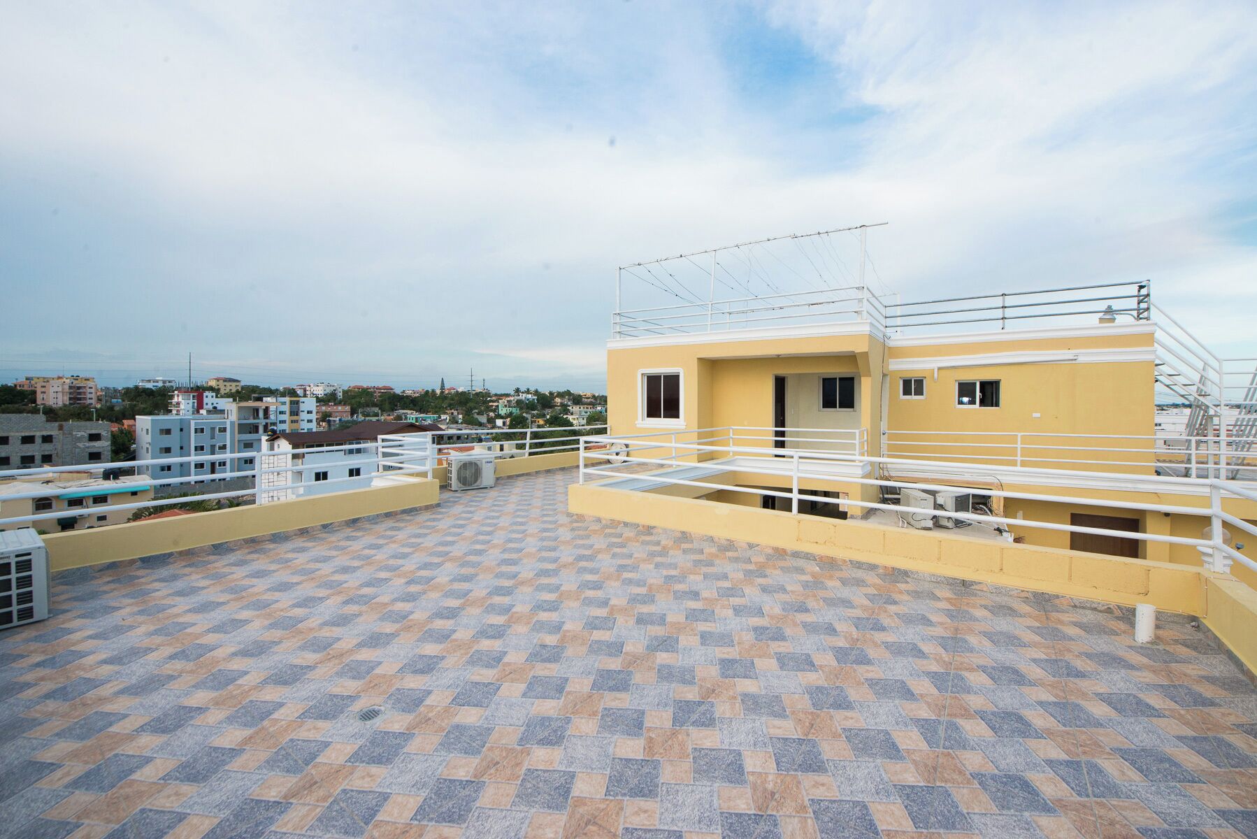 Um terraço no telhado com piso de azulejos estampados, um edifício amarelo com corrimão branco e vista para o horizonte urbano ao fundo.