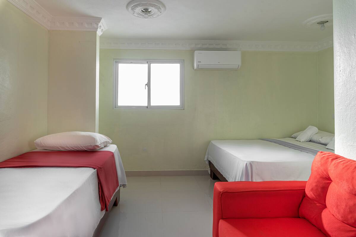 Ein kleines, gut beleuchtetes Zimmer mit zwei Einzelbetten, einem roten Sessel und einem Fenster, das natürliches Licht hereinlässt.