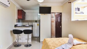 Ein kompaktes Hotelzimmer mit Bett, einer kleinen Küchenzeile, zwei Barhocker und einem an der Wand montierten Fernseher.