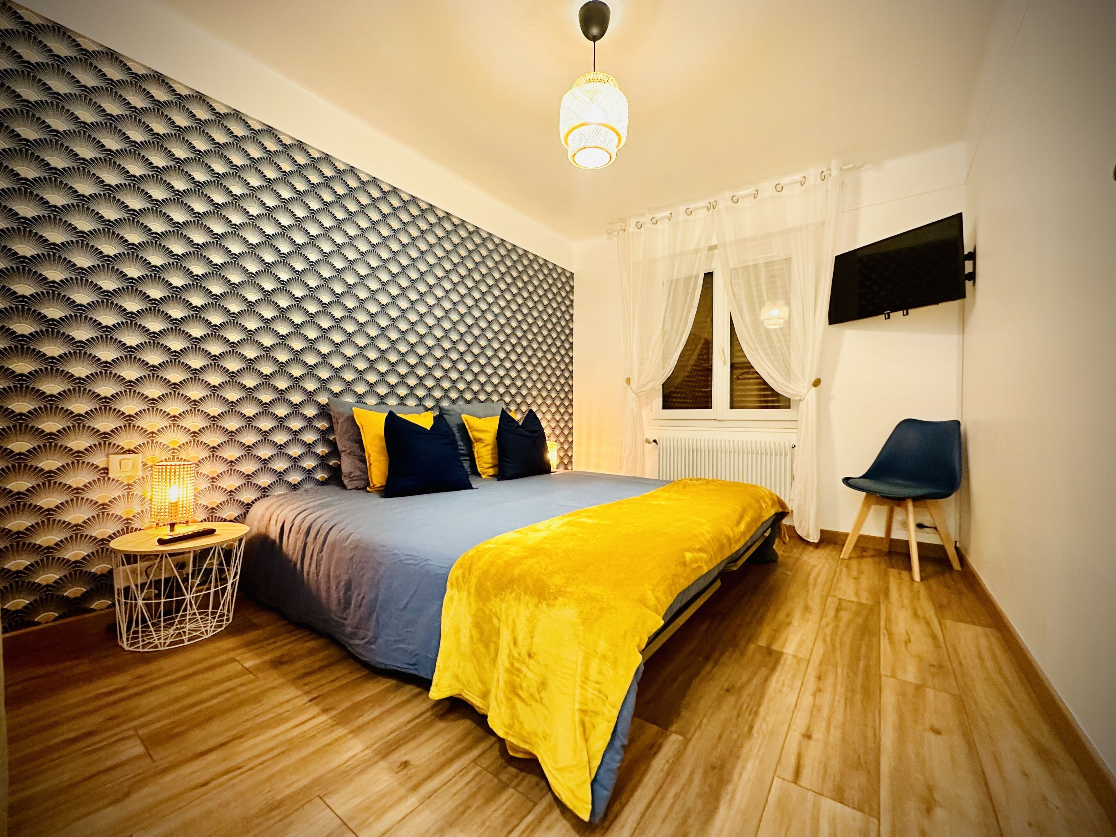 Een slaapkamer met een behang, een bed met een gele deken, een nachtkastje met een lamp, en een stoel.