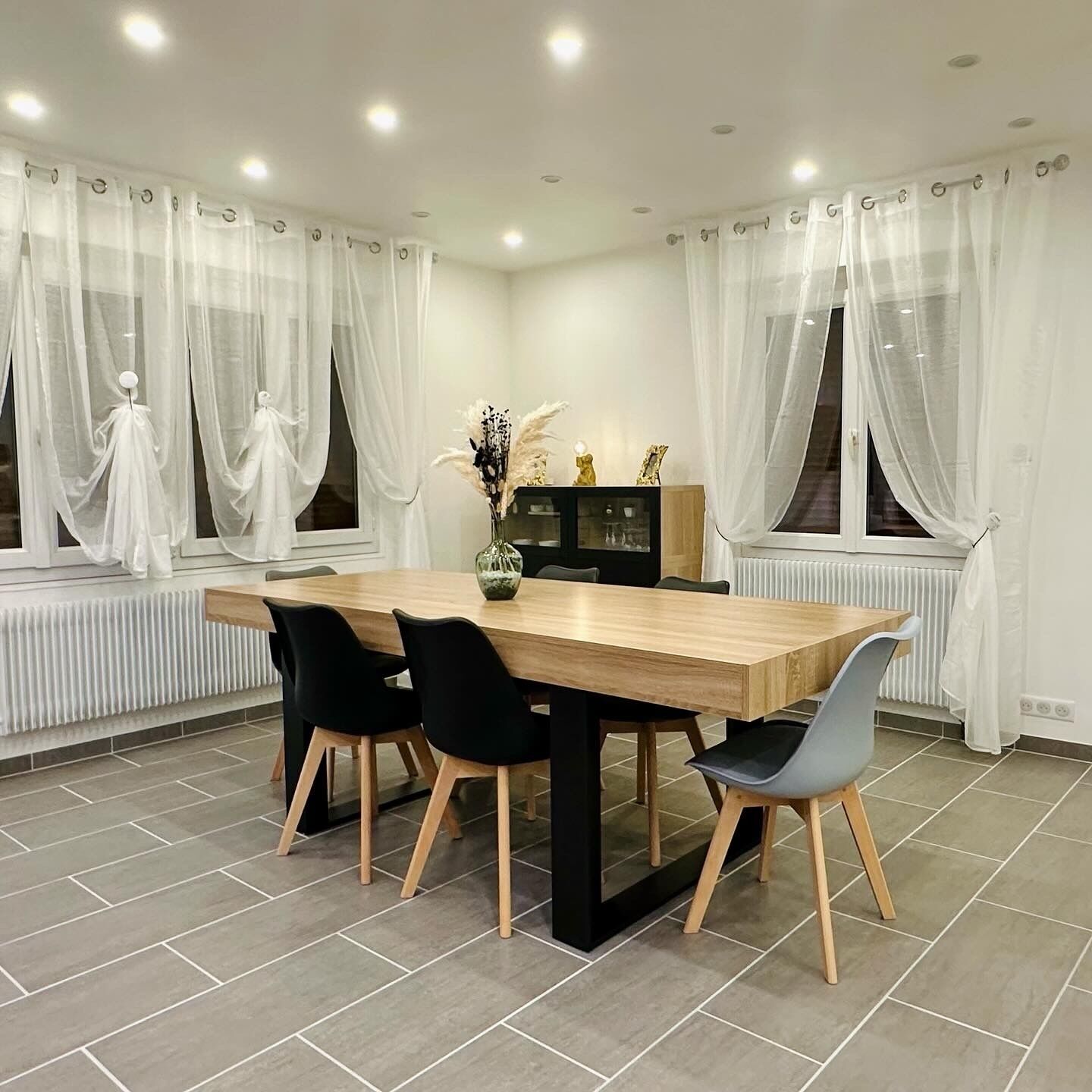 Un coin repas avec une table en bois, des chaises noires et grises, un vase avec des fleurs et de fines rideaux blancs.