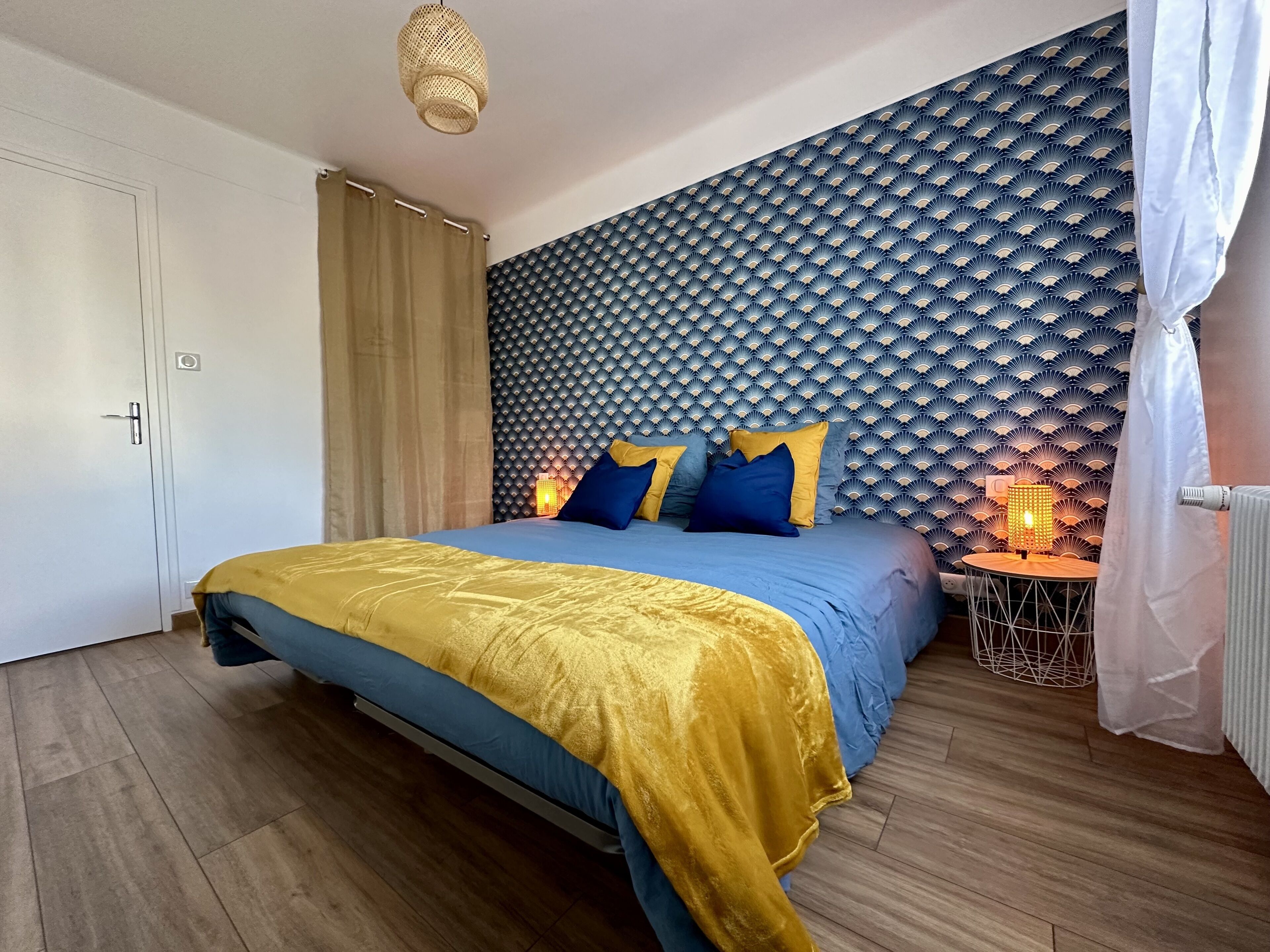 A bedroom with a bed, a headboard with a wave pattern, a bedside table with a lamp, and a curtain.